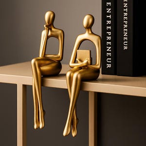 May include: Two gold abstract sculptures of seated figures. One figure rests their chin on their hand, the other holds a small book. The sculptures are placed on a gold shelf, with the word "ENTREPRENEUR" visible on a black book.
