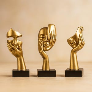 May include: Three gold-coloured face sculptures, each mounted on a black rectangular base. The sculptures depict faces with hands covering parts of them. The faces have closed eyes and are in a matte finish.