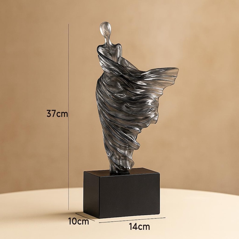 Puede incluir: Una escultura gris transl&uacute;cida de una figura con un vestido fluido, montada sobre una base rectangular negra. La escultura mide 37 cm de alto, con una base de 14 cm de ancho y 10 cm de profundidad. El vestido de la figura parece estar atrapado por el viento.