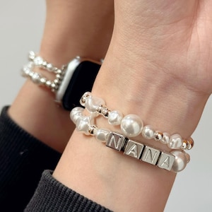 Puede incluir: Dos pulseras de perlas con detalles plateados y la palabra "NANA" escrita con cuentas de letras plateadas. Una pulsera se combina con un Apple Watch plateado. Las pulseras están en una muñeca.