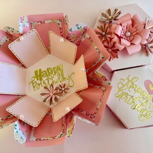 May include: Handmade pink and white hexagonal explosion boxes. The open box displays a gold script "Happy Birthday" message. The boxes are decorated with paper flowers and pearls, ideal for gifting on a special occasion.