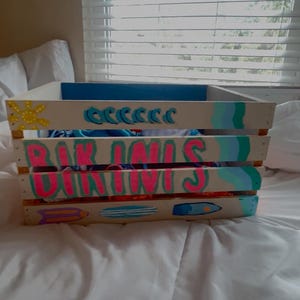 Puede incluir: Caja de madera pintada con temática playera. La caja es blanca con detalles en azul, rosa y turquesa. Presenta tablas de surf pintadas, olas y las palabras "BAR UNIS" en rosa y turquesa. Una estrella de mar y un sol amarillos también están pintados en la caja.