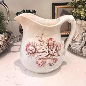 May include: A white ceramic pitcher with a floral design in pink and brown. The pitcher has a curved handle and a spout. It is sitting on a white countertop with other decorative items.