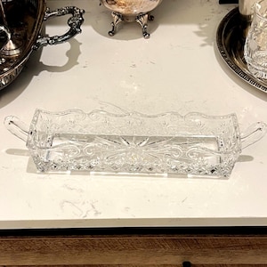 May include: A rectangular clear glass tray featuring a decorative pressed pattern with a central starburst motif and scalloped edges. The tray includes two small glass handles on opposite ends, showcasing a detailed geometric design throughout.