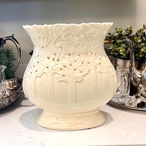 May include: Cream-colored ceramic vase with a wide, ruffled rim and a bulbous body. The vase features a repeating tree design with cut-out details. It sits on a pedestal base and is displayed on a white countertop.