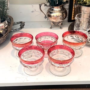 May include: Five clear glass dessert dishes with a red rim and base, arranged on a white countertop. The dishes have a decorative pattern on the base and a red interior. Other silver serving pieces and greenery are in the background.