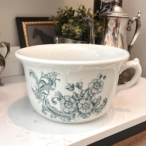 May include: A white ceramic chamber pot with a handle, decorated with teal floral and scroll patterns. The antique pot sits on a white countertop, showcasing its vintage design and decorative appeal. A classic piece for home decor.