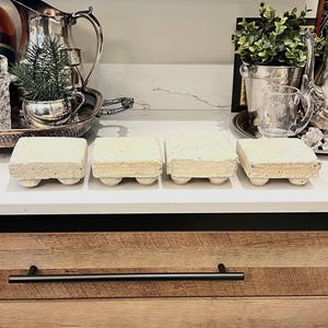 May include: Four square, off-white stone risers with a textured, porous surface. Each riser features four small, rounded feet on the bottom, providing elevation. The pieces are arranged in a horizontal row on a white countertop surface.