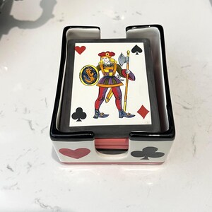 May include: A white ceramic playing card holder with black trim, featuring red heart and black club suit symbols on the front. The holder contains a deck of playing cards with a Jack of Diamonds illustration showing a figure with a shield and axe.