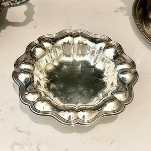 May include: A polished silver bowl featuring a scalloped, wavy edge and a smooth, reflective interior surface. The circular dish has a decorative, multi-lobed rim design that creates a distinct, undulating pattern around the entire perimeter.