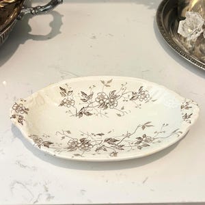 May include: An oval-shaped white ceramic dish featuring a delicate brown floral pattern with intricate leaves and blossoms across the center and edges. The dish has a slightly scalloped rim and small integrated handles on each side.