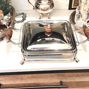 May include: A silver-plated rectangular serving dish with a clear glass lid and a wooden handle. The dish has ornate detailing and is set on a white surface. Other silver serving pieces are in the background.
