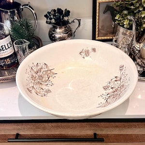 May include: A large, off-white ceramic bowl with brown floral patterns. The bowl is displayed on a white countertop, with a wooden cabinet and black hardware in the foreground. Silverware and a framed picture are in the background.