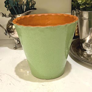 May include: A light green ceramic vase with a textured surface and a wavy, orange rim. The vase has a flared shape and sits on a white surface. Other decorative items are visible in the background.