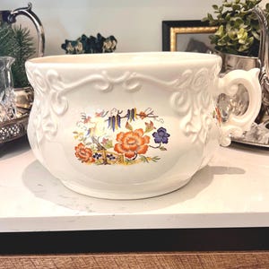 May include: White ceramic planter with ornate scrollwork and a floral design in orange, blue, and yellow. The planter has a handle and sits on a white surface. Decorative item for home decor.