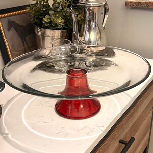 May include: A clear glass cake stand with a vibrant red base. The stand features a wide, circular glass plate supported by a flared, ruby-red pedestal. The background includes a silver tea set and a framed horse print.