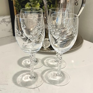 May include: Four clear crystal wine glasses with etched floral designs. The stems have a twisted rope design. The glasses are on a white surface with a silver tea set and greenery in the background.