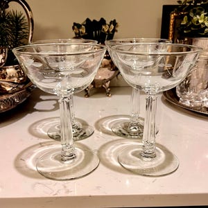 May include: Four clear champagne coupe glasses with etched floral designs. The glasses have a round base and a stem with vertical lines. The glasses are on a white surface, with other decorative items in the background.