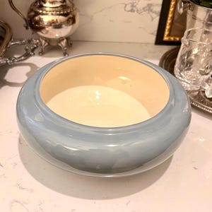 May include: A light blue ceramic bowl with a cream-colored interior. The bowl has a rounded shape and sits on a white countertop. The bowl is a decorative item.