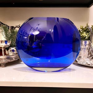 May include: A large, round, cobalt blue glass vase. The vase is spherical with a wide opening and sits on a white surface. The glass reflects the surrounding environment. Decorative home decor.