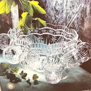 May include: A clear glass punch bowl set featuring a large scalloped bowl with starburst patterns, a matching ladle, and several small handled cups. The set includes a circular seal on the base indicating 24% lead content in the glass material.