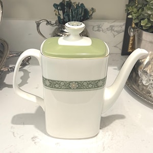 May include: White porcelain teapot with a square body, a curved handle, and a long spout. The lid is light green with a white base and a small, round knob. A floral pattern in green and white decorates the teapot.