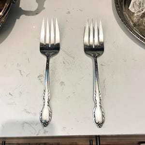 May include: Two silver-colored metal forks with four tines each. The handles feature a decorative embossed pattern with a rounded, tapered end. The finish is polished and reflective, showing a smooth surface texture along the neck and handle.