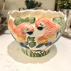 May include: A decorative ceramic bowl featuring two parrots with orange, yellow, and black details. The bowl has a scalloped rim and is decorated with green leaves and blue accents. The bowl is a decorative item.