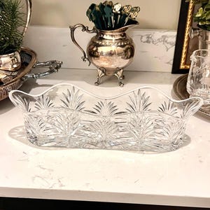 May include: A clear, rectangular crystal serving dish with a scalloped rim and a detailed fern-like pattern. The dish is designed with two handles and is placed on a white countertop. Other silver serving pieces are in the background.