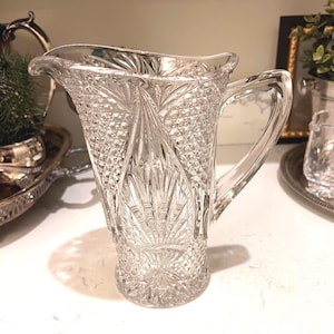 May include: A clear, cut-glass pitcher with a handle. The pitcher features a diamond and fan pattern, with a flared spout. The pitcher is sitting on a white surface. Decorative glassware.