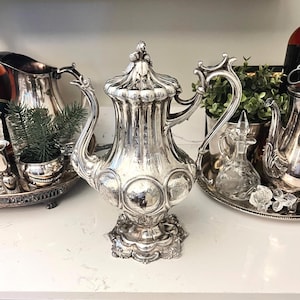 May include: A silver-plated coffee pot featuring a tall, fluted body with circular embossed medallions, a curved handle, a decorative spout, and a lid topped with a finial. The base is wide and scalloped with intricate metal detailing.