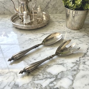 May include: A pair of silver-plated salad servers featuring elongated, ribbed handles with a decorative finial at the end. One server has a solid, rounded bowl, while the other is a three-pronged fork, both polished to a reflective, metallic finish.