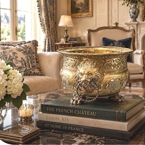 May include: A polished brass bowl featuring ornate relief carvings of lion heads with ring handles, resting on three claw feet. The bowl is placed atop a stack of three hardcover books titled The French Château, Louis XIV, and At Home in France.