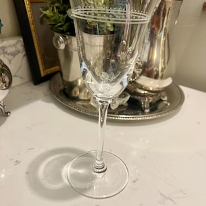 May include: A clear, stemmed wine glass with etched detailing. The glass features a delicate design around the rim and a floral motif near the base. The glass is set on a white surface, with a shadow cast below. Other silver serving pieces are in the background.