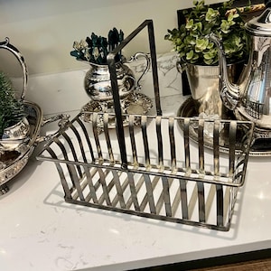 May include: A silver-toned metal basket with a rectangular shape and a handle. The basket has a lattice design on the sides and a handle that arches over the top. The basket is sitting on a white countertop.