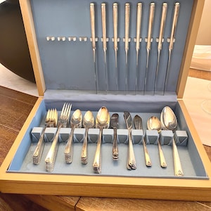 May include: A wooden silverware chest with a light blue interior houses a complete set of silver-colored flatware. The set includes forks, spoons, knives, and serving utensils, all neatly arranged. The chest is open, revealing the contents.