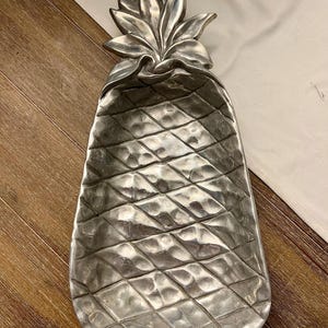 May include: A silver-colored pineapple-shaped serving tray. The tray has a textured surface with a diamond pattern and a detailed leafy top. The tray is designed to resemble a pineapple fruit.