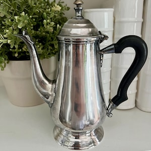 May include: A vintage silver coffee pot with a black handle and a decorative finial on the lid. The pot has a classic design with a curved spout and a round base. It is displayed on a white shelf.