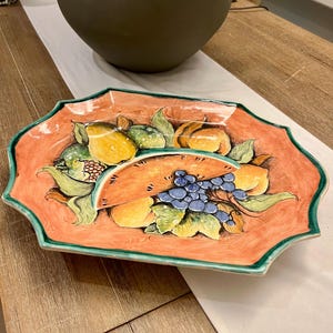 May include: A rectangular ceramic serving platter with a scalloped edge. The platter features a vibrant orange background with a painted design of various fruits, including lemons, grapes, and pumpkins. The edge is trimmed with a green border.