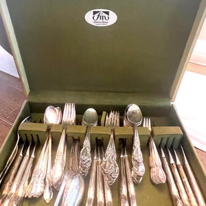 May include: An open, green velvet-lined silverware case filled with an assortment of silver spoons, forks, and knives. The silverware has ornate handles. The case has a white sticker with black text.