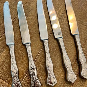 May include: Five silver butter knives with ornate handles featuring a grape and leaf design. The blades are smooth and reflect light. The handles have a decorative initial. The knives are arranged on a wooden surface.