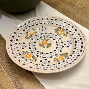 May include: A decorative ceramic plate with a floral design. The plate features a cream-colored base with a blue and yellow floral pattern. The rim is decorated with a repeating blue pattern. The plate is placed on a white cloth.