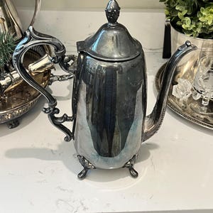 May include: A polished silver-toned metal teapot featuring a tall, cylindrical body, a curved handle with ornate scrollwork, a long slender spout, and a domed lid topped with a small finial. The teapot stands on three small, decorative feet.