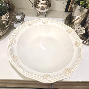 May include: A white, decorative serving bowl with an ornate, scalloped edge. The rim features a raised, textured design with shell-like accents. The bowl is empty and sits on a white surface, suggesting a kitchen or dining setting.