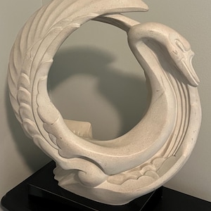 May include: A cream-colored swan sculpture in a circular design. The swan's body curves to form a circle, with its head and neck reaching towards its back. The sculpture has detailed feather and wing patterns, and sits on a black surface.