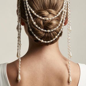 May include: A bridal hairstyle with a braided bun, embellished with multiple strings of pearls and clear beads. The pearl strands fall down the back, with teardrop-shaped pearl drops. The woman is wearing a white dress.