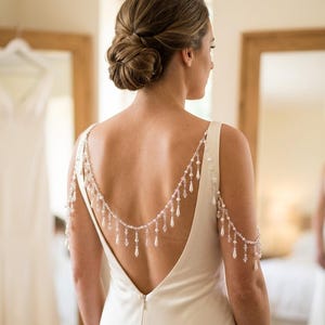 May include: A back necklace accessory featuring silver-toned chains draped across the shoulders and back, adorned with small white pearls and clear teardrop-shaped crystals that hang in a fringe pattern, attached to a sleeveless white dress.