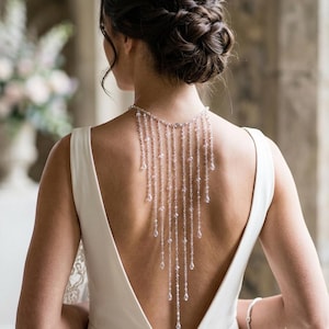 May include: A silver-toned back necklace featuring multiple cascading chains adorned with clear, teardrop-shaped crystals. The jewelry piece attaches around the neck and drapes down the back, worn over a sleeveless, solid white dress with a deep scoop back.