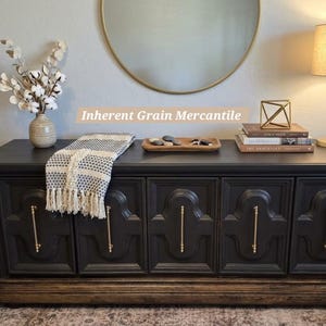 May include: A black wooden storage chest with five cabinet doors, each featuring a vertical gold handle. The chest has a dark wood base and is topped with a white and black patterned throw blanket, a wooden tray with stones, and books.