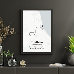 May include: A framed print featuring a black and white map of a triathlon course in Emilia Romagna. The word "Triathlon" is displayed in a bold font, with the region's name below. The print includes icons representing the distances of the swim, bike, and run.